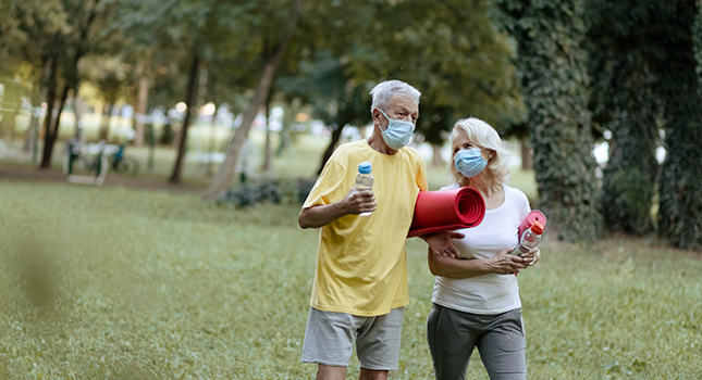 Older couple walking outside with masks and man is holding a yoga mat 